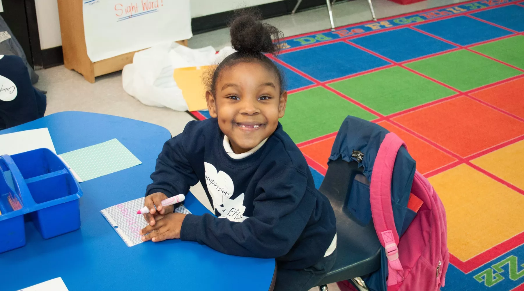 A young girl with a ponytail missing her two front teeth smiling big at the camera while sitting at her desk writing with a pink marker