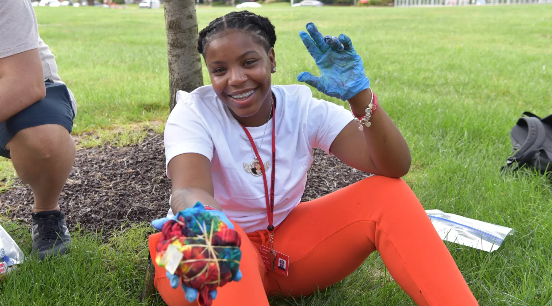 TILE student holding out tie-dyed shirt and holding up "L" hand symbol while sitting in grass on University of Louisville campus in front of the College of Business.