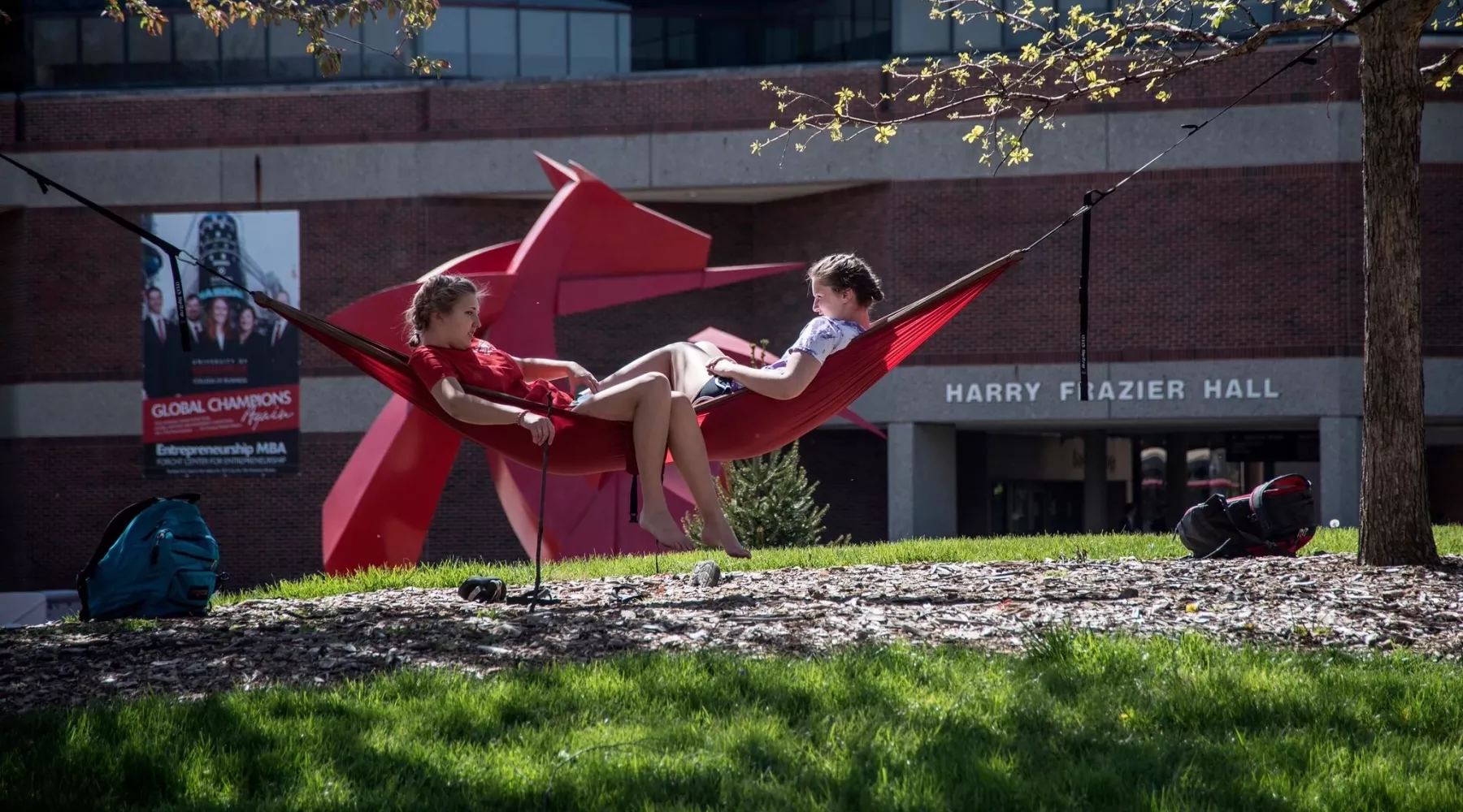 Two female students sharing a hammock outside of the College of Business with Big Red in the background