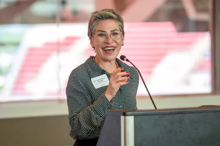 UofL College of Business MBA Program director, Anne Zwerg,  gives an opening speech at a podium of the PNC club. A window is in the background that look out onto the stands of Cardinal Stadium.