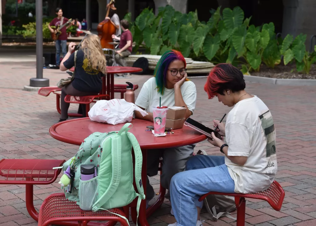 Student with rainbow hair watching red haired student write on tablet while sitting outside the SAC at a picnic table