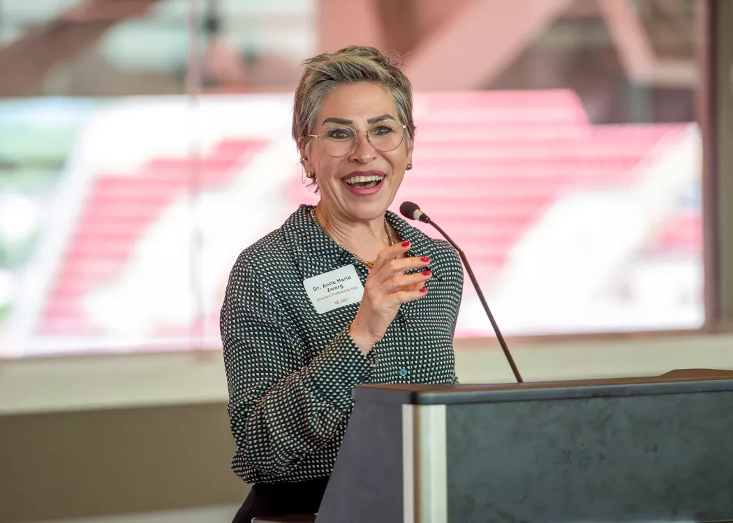 UofL College of Business MBA Program director, Anne Zwerg,  gives an opening speech at a podium of the PNC club. A window is in the background that look out onto the stands of Cardinal Stadium.