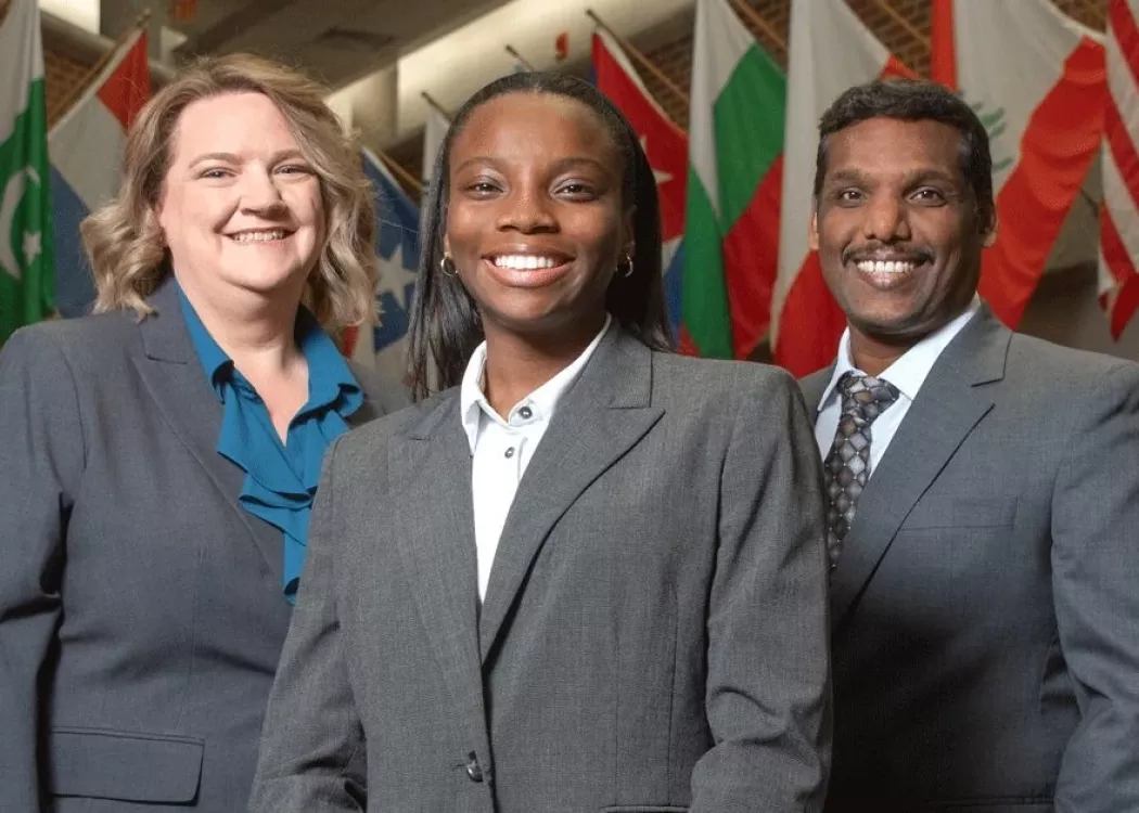 Three suited graduate students together in front of a row of flags.