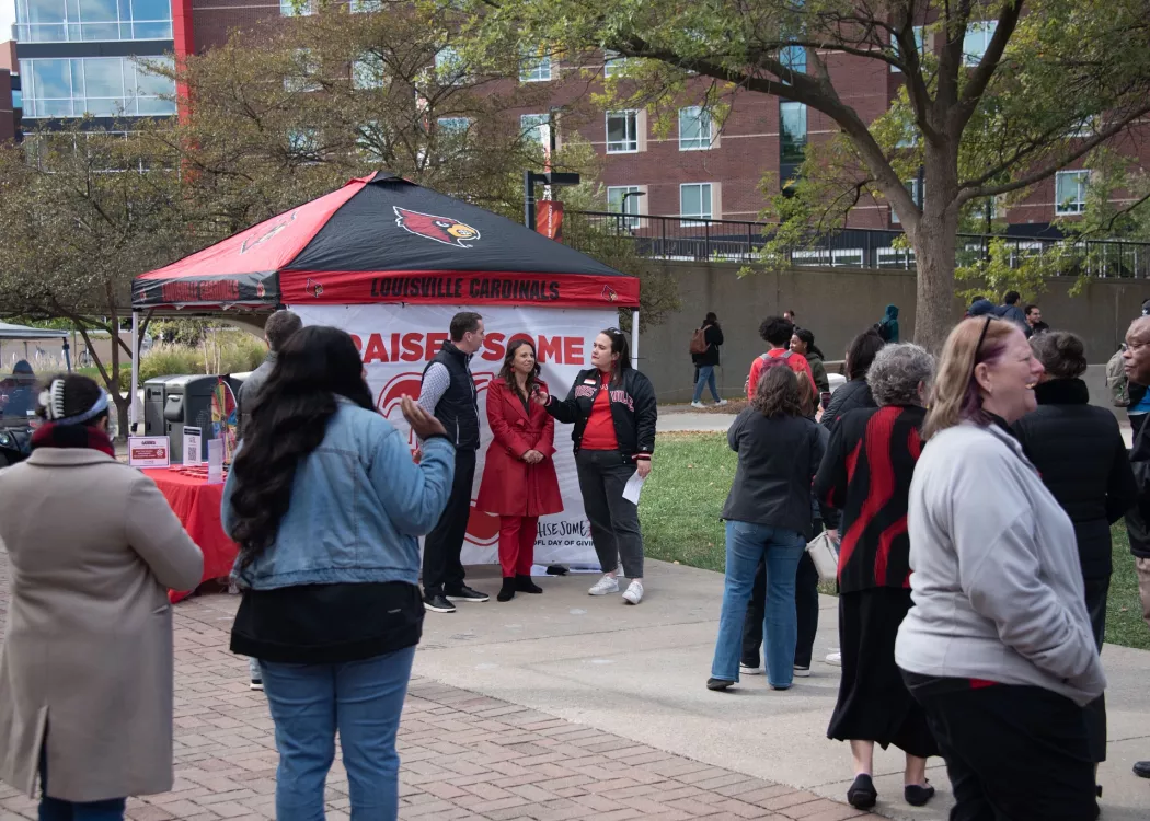 A crowd gathers around a woman in a University of Louisville Jacket interviewing two people.