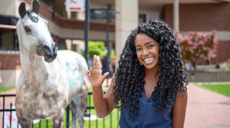 Student with L's up in front of horse statue by the College of Business