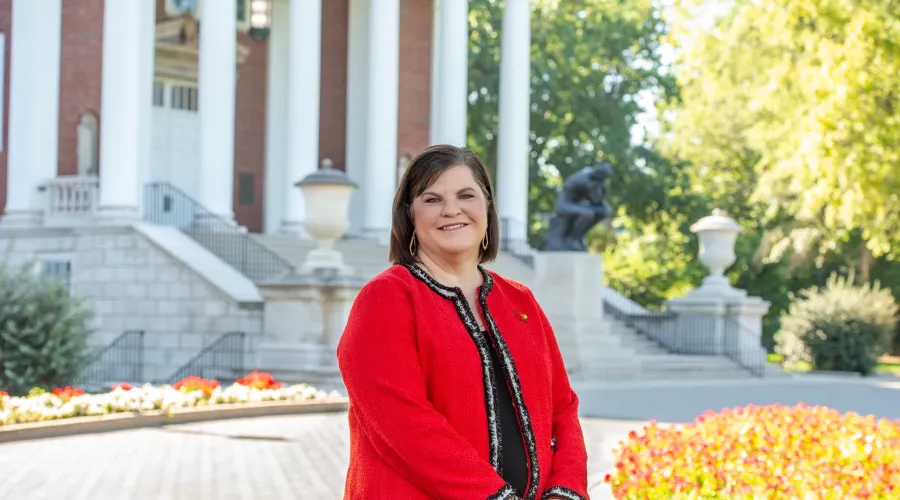 Woman in Cardinal red blazer standing in front of building with columns and bronze statue of The Thinker on UofL's Belknap…