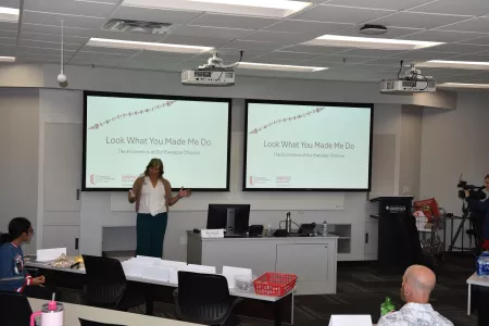 Beth Munnich standing in front of a presentation that reads "Look what you made me do. The economics of our everyday…